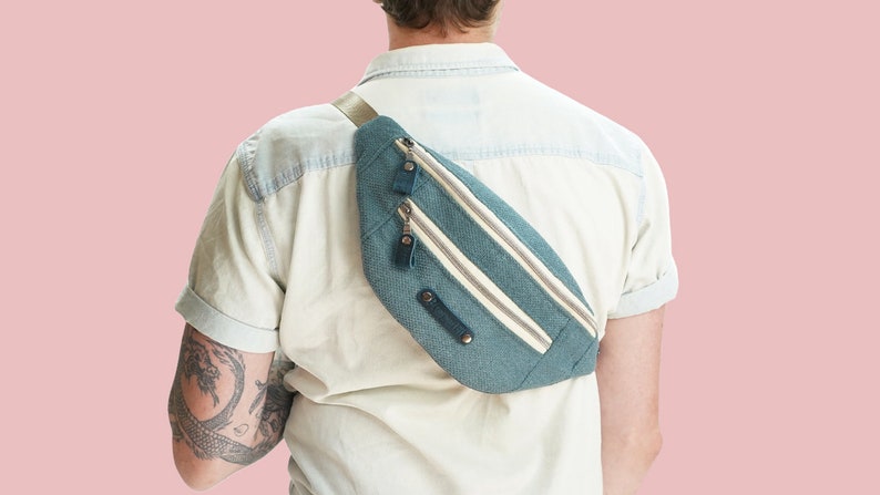 K&ouml;nnte beinhalten: Eine t&uuml;rkisfarbene Bauchtasche mit wei&szlig;en Streifen und zwei Rei&szlig;verschl&uuml;ssen. Die Bauchtasche wird &uuml;ber dem K&ouml;rper einer Person getragen, die ein hellblaues Hemd tr&auml;gt.