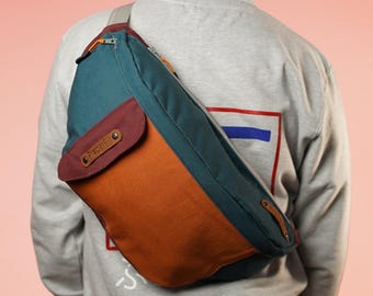 May include: A teal, brown, and burgundy crossbody bag with a leather patch that says "Front" on it. The bag is worn over a grey sweatshirt.