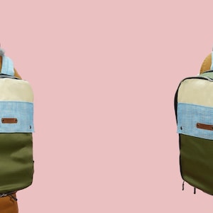 Pode incluir: Duas mochilas com um design de blocos de cores s&atilde;o mostradas por tr&aacute;s. As mochilas t&ecirc;m uma parte superior creme, um centro azul claro e uma parte inferior verde oliva. As al&ccedil;as s&atilde;o azuis claras e as mochilas t&ecirc;m uma etiqueta de couro castanho.