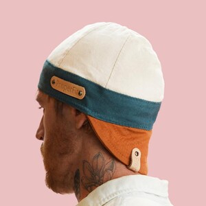 May include: A two-tone cap with a cream-colored top and a teal band. The cap has a brown leather tag with the text "ProperFit". The lower portion is orange, with a leather strap and button closure.