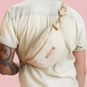 Puede incluir: Una riñonera blanca de piel sintética con una cadena dorada y una correa de cuero marrón. La bolsa se lleva cruzada en el cuerpo de una persona que lleva una camisa blanca.