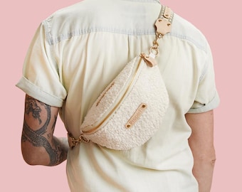 May include: A white faux fur fanny pack with a gold chain and a brown leather strap. The bag is worn across the body of a person wearing a white shirt.