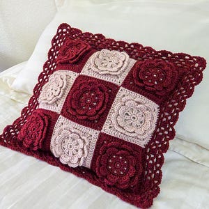 Może przedstawiać: Kwadratowa, szydełkowa poduszka z kwiatowym wzorem w odcieniach bordowego i beżowego. Poduszka ma falisty brzeg i jest umieszczona na białym łóżku. W tle mały stolik nocny z lampą i rośliną doniczkową.