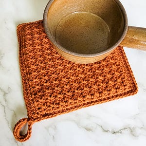 Rust-colored Classic Potholder crocheted in dense, double-thick Trinity Stitch fabric with a hanging loop. A clay bowl rests on top of the potholder, which is displayed on a white marble countertop.