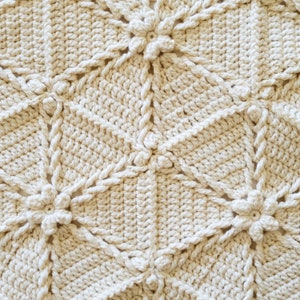 Close-up of a crocheted blanket with a repeating pattern of flowers. The blanket is made of a cream-colored yarn.