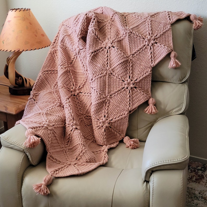 A soft, blush pink crocheted throw blanket with a braided starburst pattern and tassels. The blanket is draped over a light beige armchair.