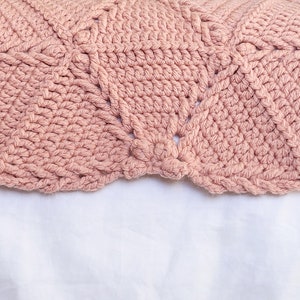 A close-up of a crocheted blanket in a dusty rose color. The blanket has a repeating pattern of diamond shapes with a flower-like design in the center of each diamond.