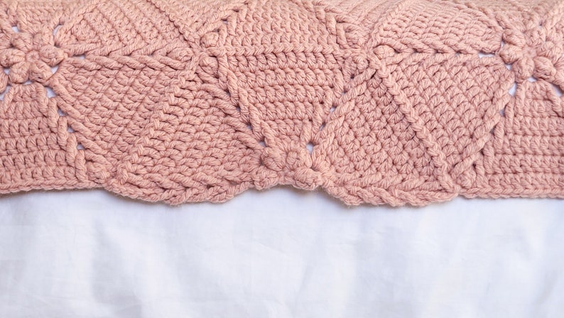 A close-up of a crocheted blanket in a dusty rose color. The blanket has a repeating pattern of diamond shapes with a flower-like design in the center of each diamond.