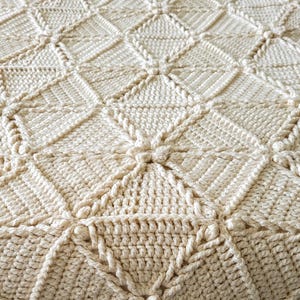 A crocheted throw blanket in a cream color with a diamond pattern, braided texture, and tassels.
