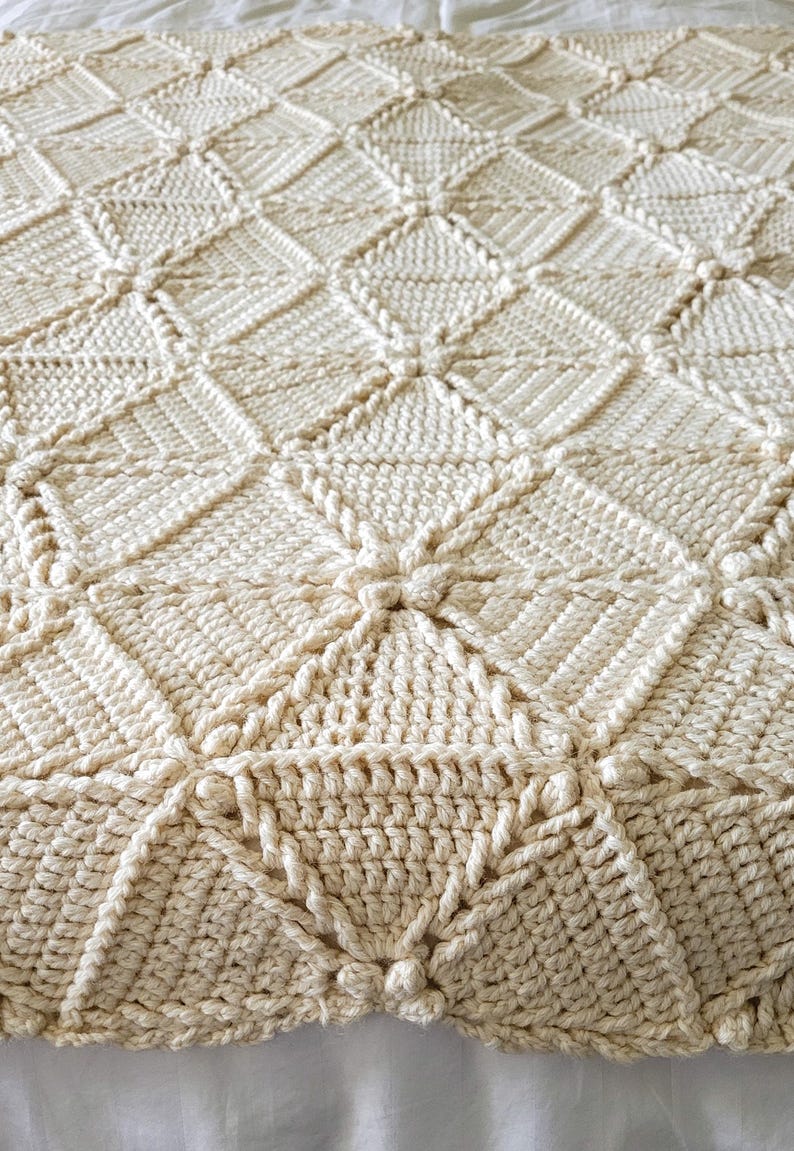 A crocheted throw blanket in a cream color with a diamond pattern, braided texture, and tassels.