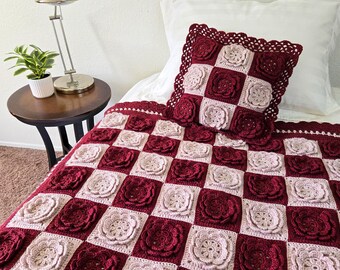 May include: A handmade crochet blanket and pillow set. The blanket and pillow feature a checkerboard pattern of burgundy and light pink squares, each with a raised floral design. The pillow has a decorative burgundy border.