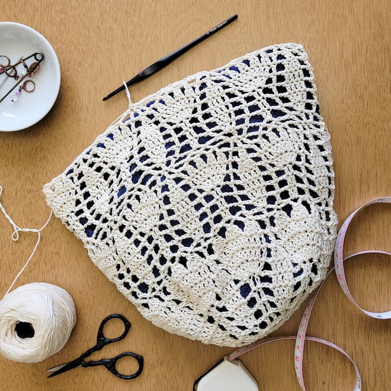 A white crocheted lace produce bag in progress. A dark blue fabric placed inside shows off the lace stitches. The bag has a drawstring closure and the lace design features bell-shaped flowers and leaves. The bag is lying on a wooden surface with a measuring tape, crochet hook, yarn, and scissors.