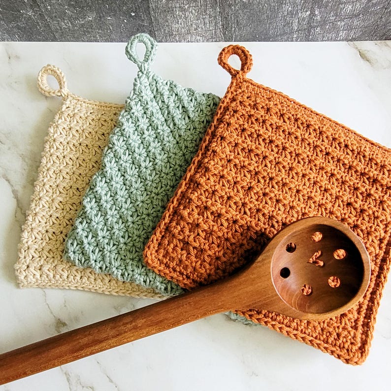 Three double-thick crochet potholders in beige, sage green, and rust are displayed on a white marble countertop beside a wooden spoon. Each features textured Trinity Stitch fabric and a hanging loop.
