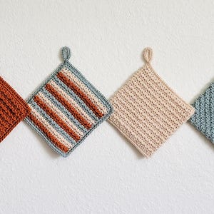 Four double-thick Trinity Stitch potholders crocheted in cotton yarn are hung on a white wall. The potholders are shown in sage green, beige, rust, and a three-color striped version, each featuring dense, highly textured star-stitch fabric.