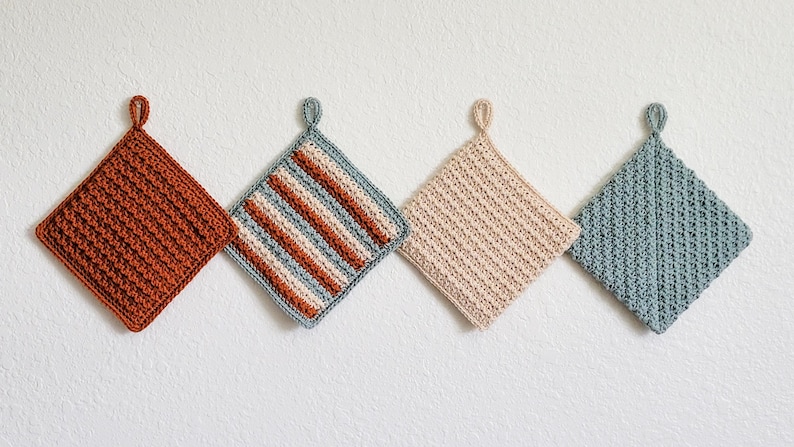 Four double-thick Trinity Stitch potholders crocheted in cotton yarn are hung on a white wall. The potholders are shown in sage green, beige, rust, and a three-color striped version, each featuring dense, highly textured star-stitch fabric.