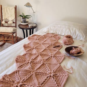 A pink crocheted blanket made of hexagonal shapes with a flower design in the center of each hexagon. The blanket is partially assembled with some of the hexagons still separate. A white throw with tassels is draped over the rocking chair in the background.