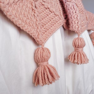 A pink crocheted blanket with three tassels. The blanket is draped over a white bedspread.