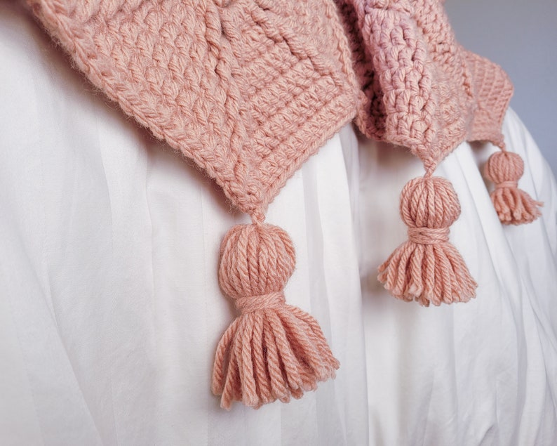 A pink crocheted blanket with three tassels. The blanket is draped over a white bedspread.