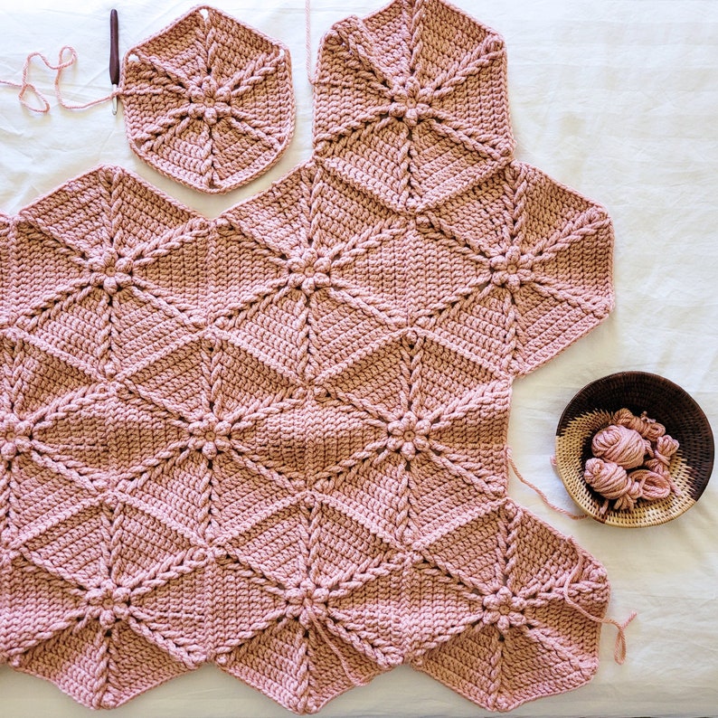 A pink crocheted blanket made of hexagonal shapes with a flower design in the center of each hexagon. The blanket is partially assembled with some of the hexagons still separate.