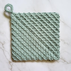 Sage green Folded Potholder crocheted in dense, double-thick Trinity Stitch fabric with a hanging loop. The folded construction creates a distinctive diagonal seam across the front. The potholder is displayed on a white marble countertop.