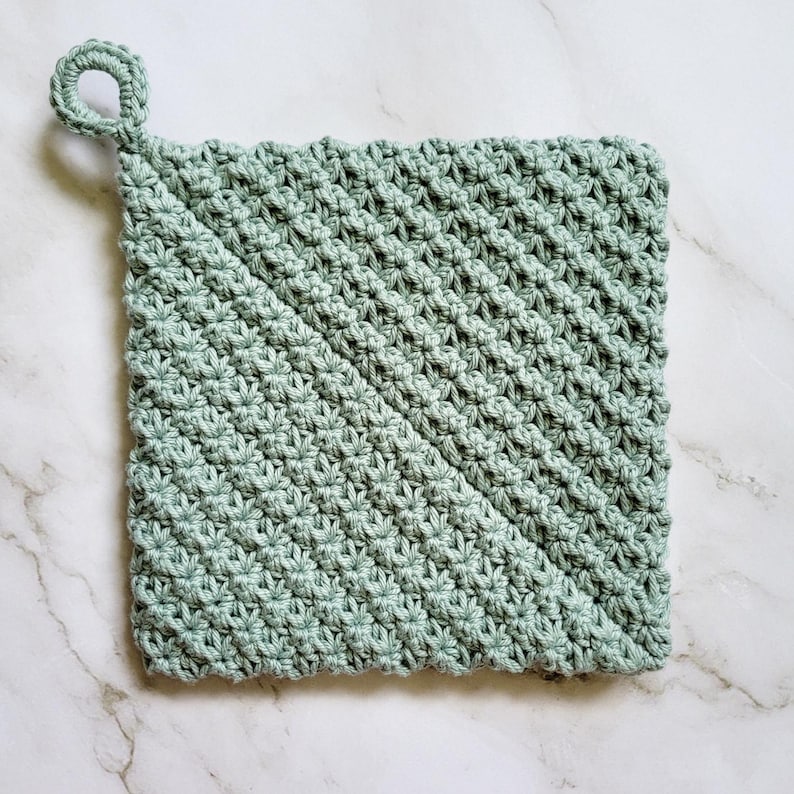 Sage green Folded Potholder crocheted in dense, double-thick Trinity Stitch fabric with a hanging loop. The folded construction creates a distinctive diagonal seam across the front. The potholder is displayed on a white marble countertop.