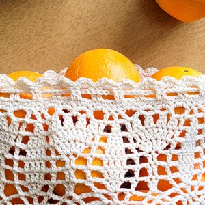 A closeup of a white crocheted produce bag filled with oranges. The bag has a drawstring closure and the lace design features bell-shaped flowers and leaves.