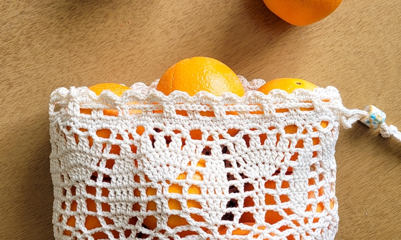 A closeup of a white crocheted produce bag filled with oranges. The bag has a drawstring closure and the lace design features bell-shaped flowers and leaves.