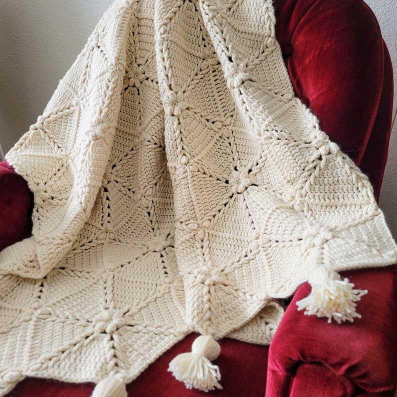 A crocheted throw blanket in a cream color with a diamond pattern and tassels. The blanket is draped over a red armchair.