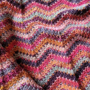 Op de afbeelding: Close-up van een handgemaakte gehaakte deken met een chevronpatroon. De deken heeft een levendige mix van kleuren, waaronder roze, oranje, paars en blauwgroen. De getextureerde steken creëren een gezellig en visueel aantrekkelijk ontwerp.