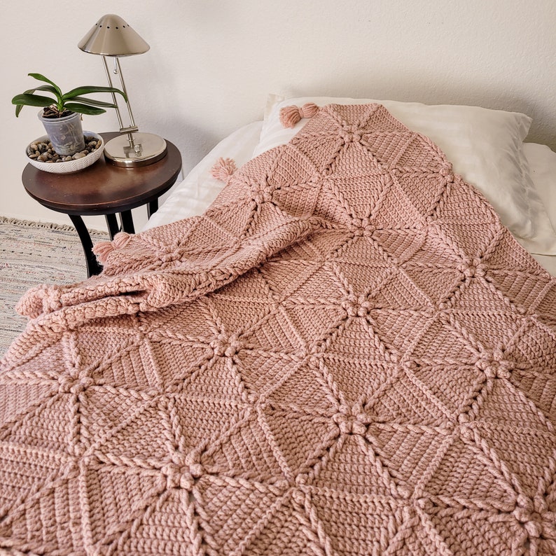 A blush pink crocheted throw blanket with a braided pattern. The blanket is folded and draped over a white bed with a white pillow.