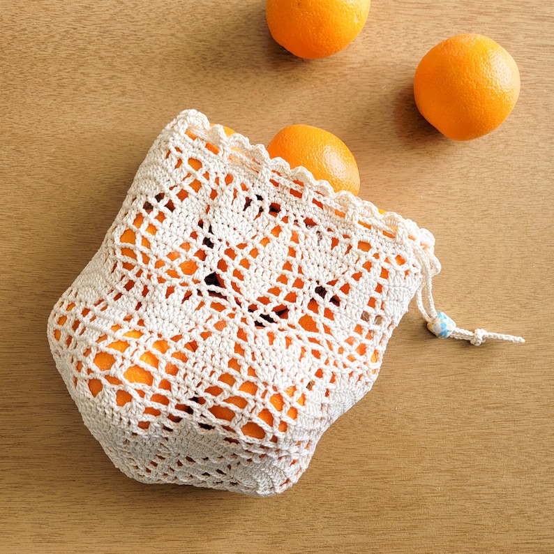 A white crocheted lace produce bag filled with oranges. The bag has a drawstring closure and the lace design features bell-shaped flowers and leaves.