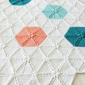 A white crocheted blanket with a geometric pattern featuring teal, light blue, and coral hexagons.
