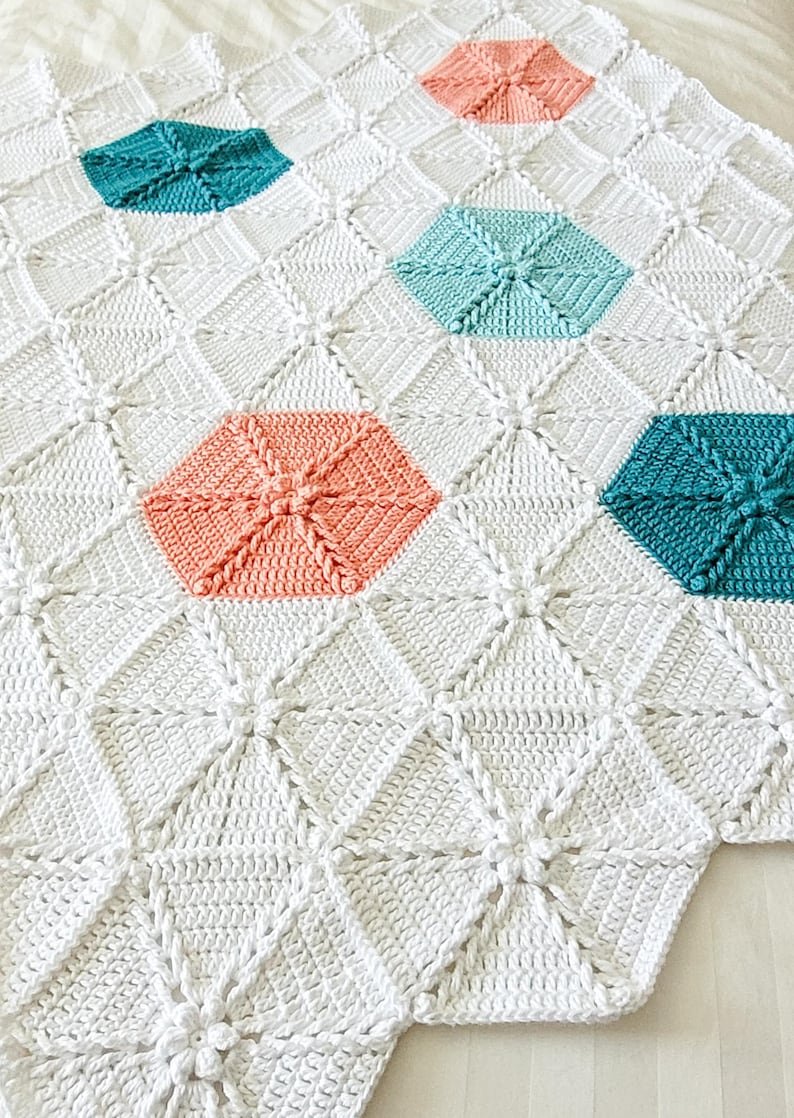 A white crocheted blanket with a geometric pattern featuring teal, light blue, and coral hexagons.