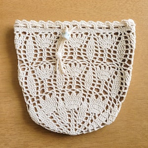 A white crocheted lace produce bag lays flat on a wooden table. The bag has a drawstring closure and the lace design features bell-shaped flowers and leaves.