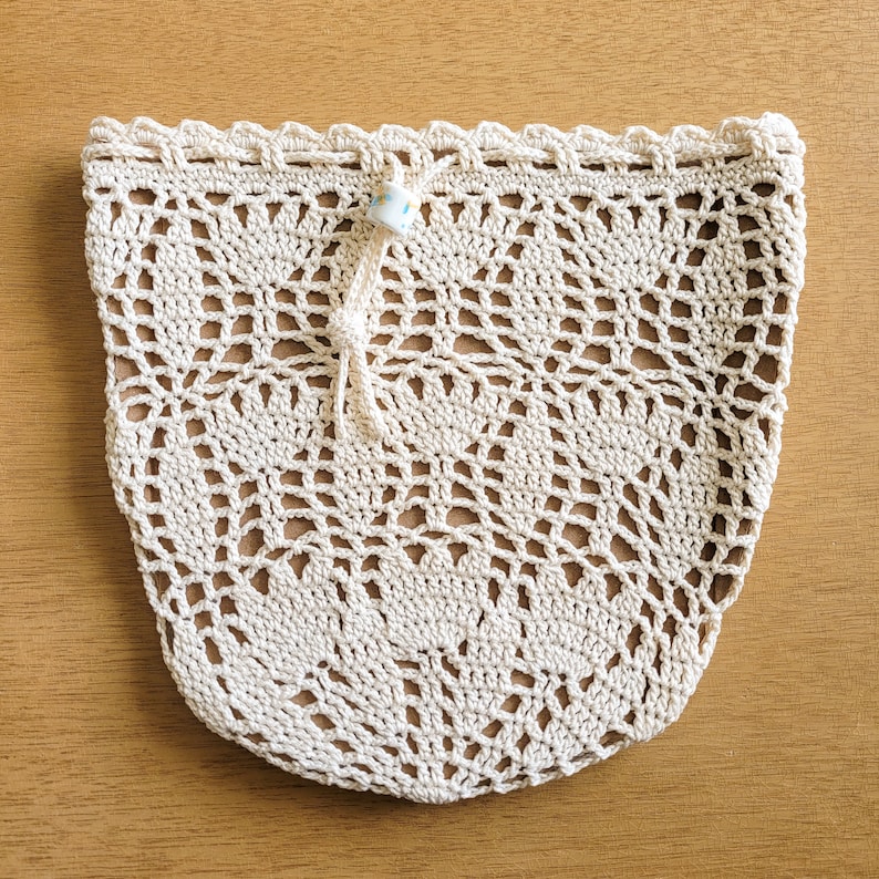 A white crocheted lace produce bag lays flat on a wooden table. The bag has a drawstring closure and the lace design features bell-shaped flowers and leaves.