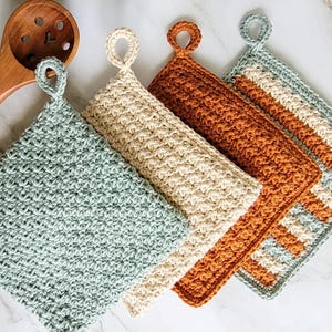 Peut inclure: Quatre maniques carrées en crochet avec des boucles de suspension, présentant des motifs de points texturés en vert sauge, crème et orange brûlé, disposées en diagonale à côté d'une cuillère à fentes en bois.