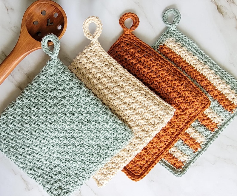Four double-thick Trinity Stitch potholders crocheted in cotton yarn are arranged on a white marble countertop beside a wooden spoon. The potholders are shown in sage green, beige, rust, and a three-color striped version, each featuring dense, highly textured fabric with a flower- or star-like appearance and a hanging loop.