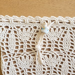 A close-up of a white crocheted lace produce bag. The bag has a drawstring closure and the lace design features bell-shaped flowers and leaves.