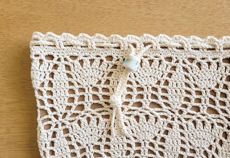 A close-up of a white crocheted lace produce bag. The bag has a drawstring closure and the lace design features bell-shaped flowers and leaves.