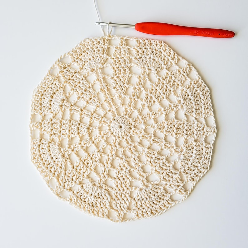 A crocheted lace circle stitched with a white thread, showing the first twelve rounds of the Bellflower Produce Bag. A metal crochet hook with a red handle is at the top of the photo.