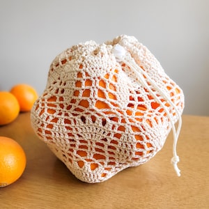 A white crocheted produce bag filled with oranges. The bag has a drawstring closure and the lace design features bell-shaped flowers and leaves.
