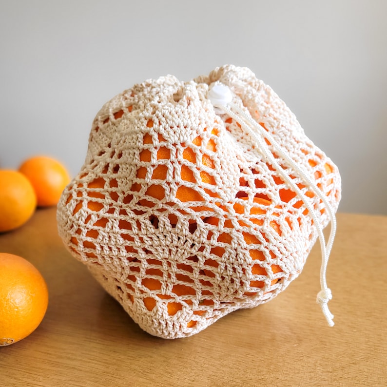 A white crocheted produce bag filled with oranges. The bag has a drawstring closure and the lace design features bell-shaped flowers and leaves.