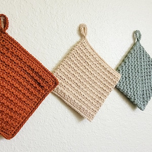 Three double-thick Trinity Stitch potholders crocheted in cotton yarn are hung on a white wall. The potholders are shown in sage green, beige, rust, and a three-color striped version, each featuring dense, highly textured star-stitch fabric. Photo is taken at an angle to highlight the textured stitch pattern.