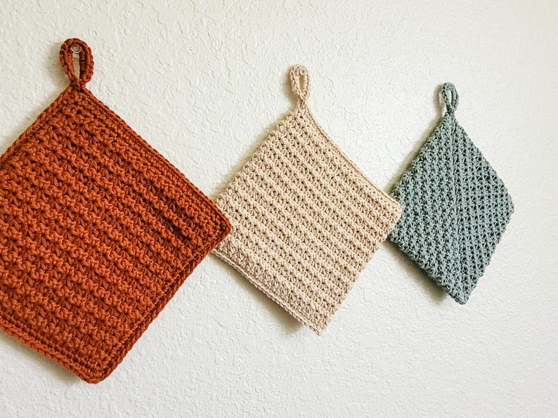 Three double-thick Trinity Stitch potholders crocheted in cotton yarn are hung on a white wall. The potholders are shown in sage green, beige, rust, and a three-color striped version, each featuring dense, highly textured star-stitch fabric. Photo is taken at an angle to highlight the textured stitch pattern.