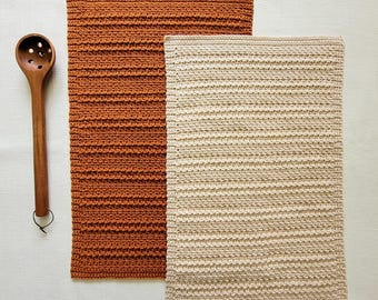 May include: Two rectangular crochet dishcloths, one in a deep burnt orange and one in a light cream color, featuring a textured horizontal ribbed pattern. A wooden slotted spoon with a long handle is positioned to the left of the cloths.