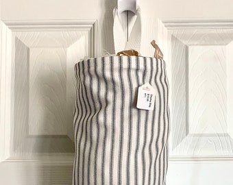 May include: A vertically striped fabric bag, primarily white with dark gray stripes, hangs from a white hook on a white door. The bag is cylindrical with a gathered bottom. A small tag is attached near the top.
