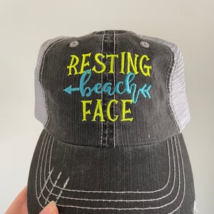 May include: A black and gray trucker hat with a distressed look. The hat has a white mesh back and a yellow embroidered design that says "Resting Beach Face" with a blue arrow pointing to the right.