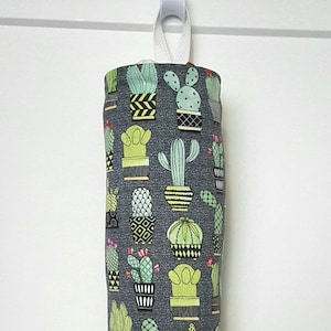 May include: A fabric bag with a white loop for hanging, patterned with various green and yellow cacti on a dark grey background. The bag is cylindrical and gathered at the bottom.