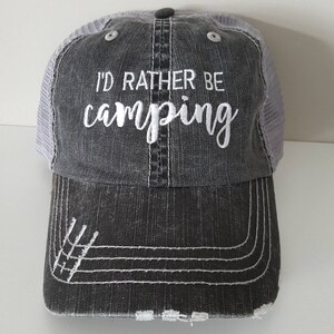 Peut inclure: Un chapeau de camionneur gris et blanc à l'aspect vieilli. Le chapeau a un dos en maille blanche et un devant noir avec le texte "I'd rather be camping" brodé en blanc.