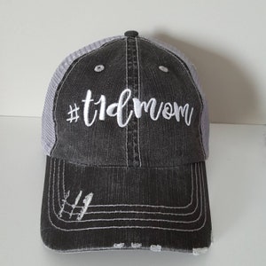May include: A black and gray distressed baseball cap with a white mesh back. The front of the cap has white embroidery that reads "#tidmom".